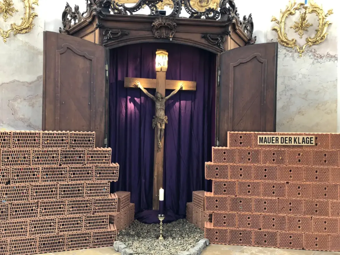 Ein Kreuz mit Jesusfigur, umgeben von Ziegelmauern und lila Vorhängen. Ein Kerzenlicht lädt zum Kreuz Gebet ein.