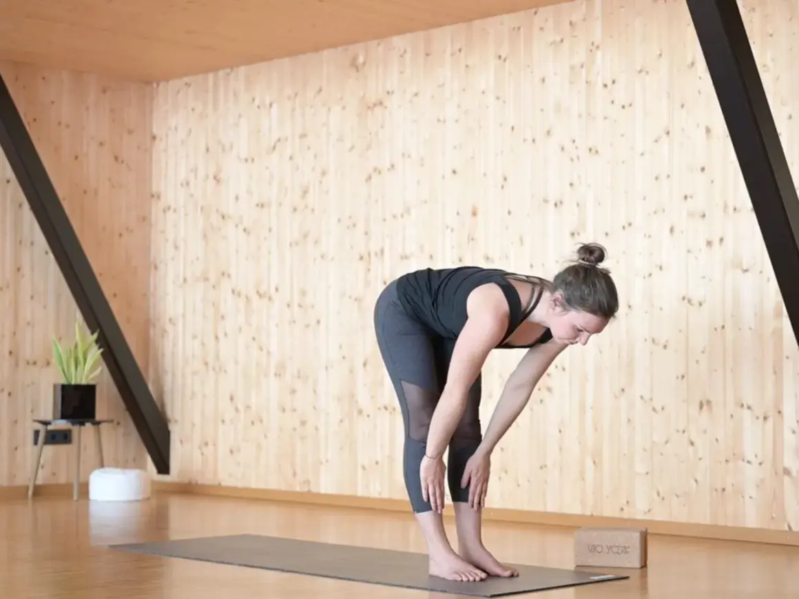 Frau in Uttanasana, einer Vorwärtsbeuge im Surya Namaskar Yoga, dehnt sich auf einer Matte.