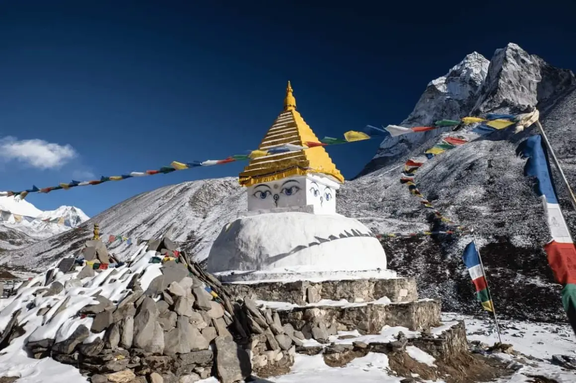 Ein buddhistischer Stupa mit Augen im Schnee, umgeben von Gebetsfahnen, die buddhistische Mantras in den Wind tragen. Majestätische Berge im Hintergrund.