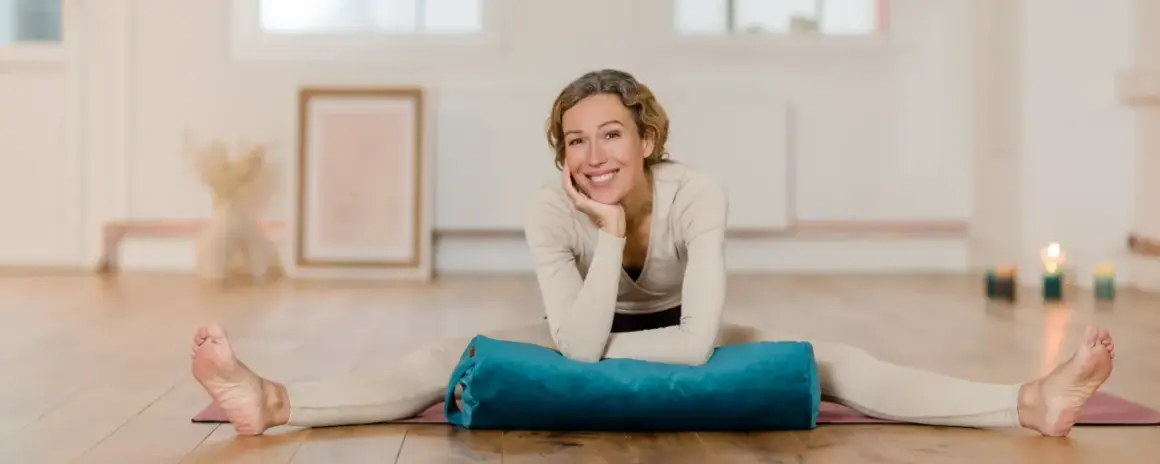Frau lächelt in einem hellen Raum, Beine gespreizt, mit einem blauen Bolster für Nadi Yoga.