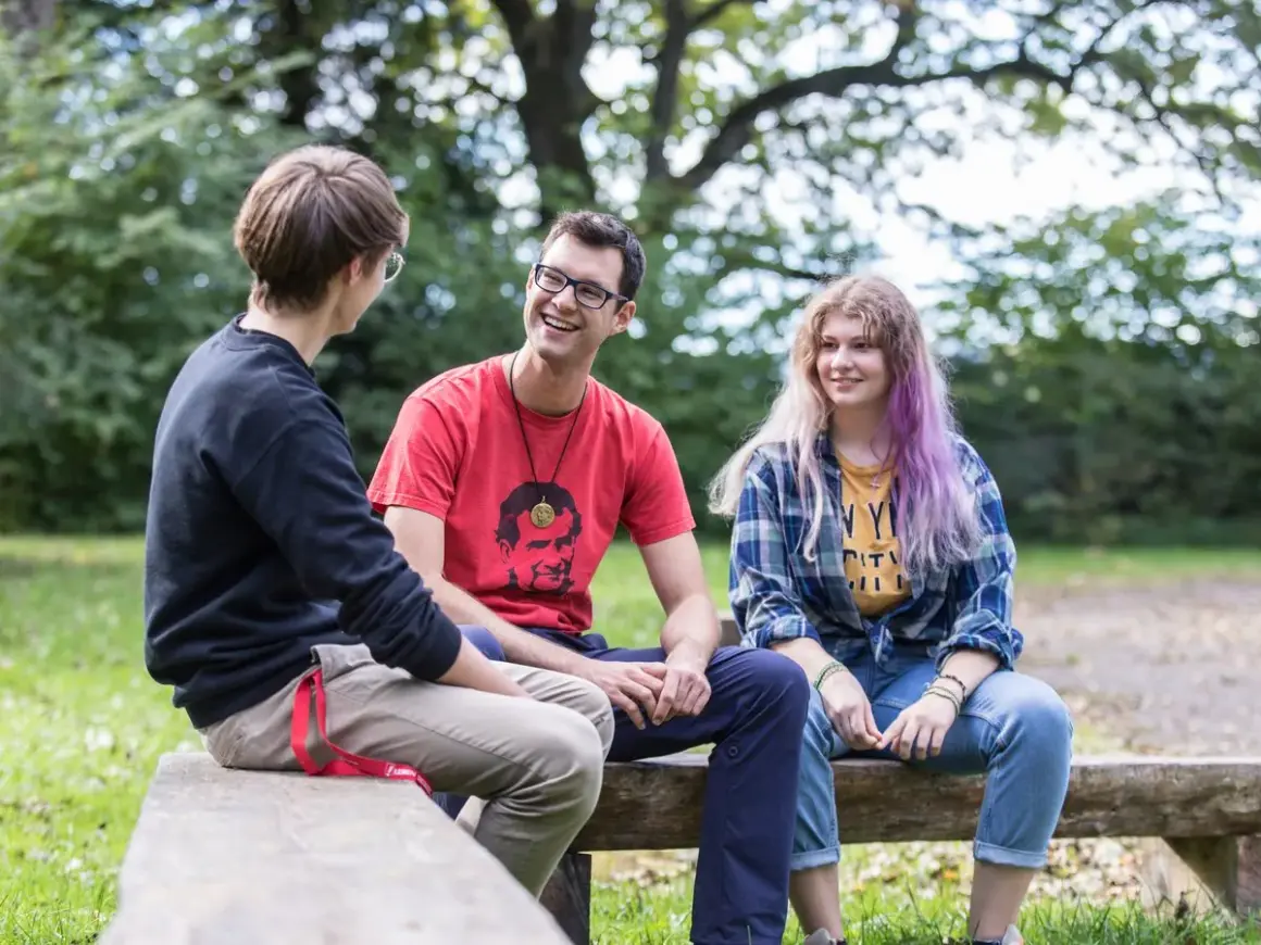 Drei junge Leute unterhalten sich lachend auf einer Bank im Freien. Der Mann in Rot trägt ein T-Shirt mit einem Porträt, das an den **Salesianer Orden** erinnert.