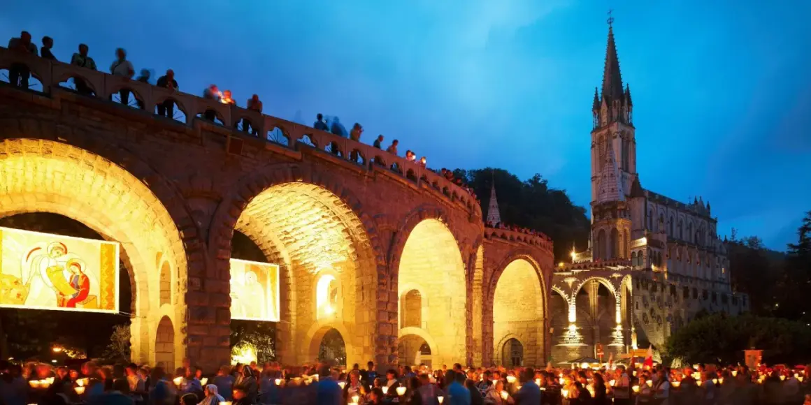 Eine Lichterprozession in Lourdes. Menschen halten Kerzen unter einem steinernen Bogen, während im Hintergrund eine Kirche beleuchtet ist.