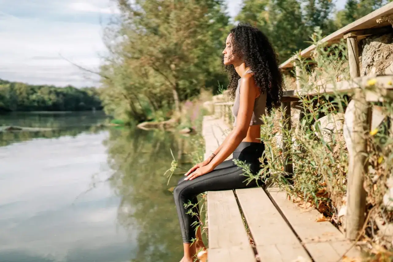 Frau meditiert am Wasser. Sie sitzt auf einer Holzplattform, die Augen geschlossen, und genießt die Ruhe. So kann man meditieren.