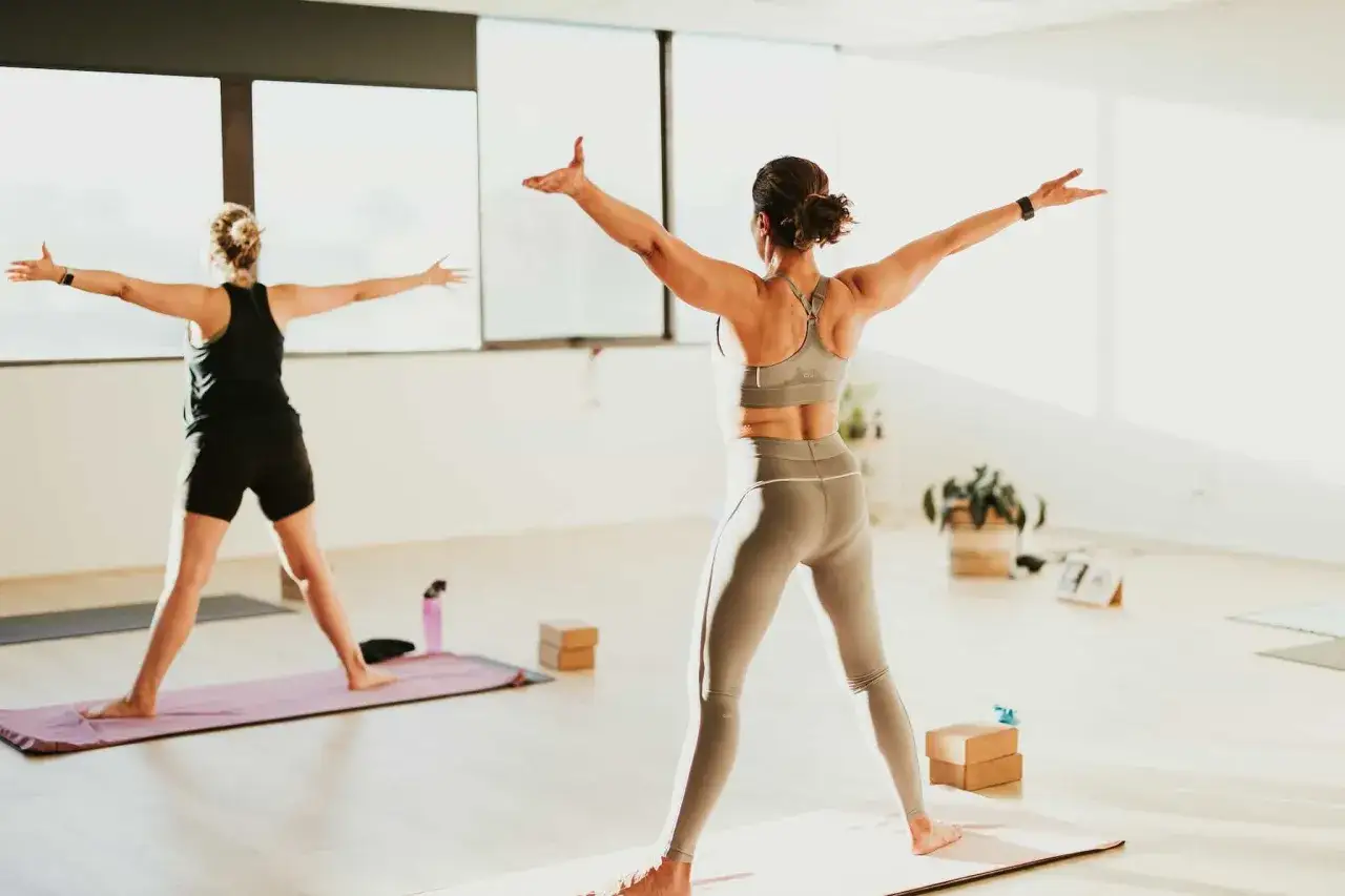 Zwei Frauen praktizieren Bikram Hot Yoga in einem hellen Raum. Sie stehen auf Yogamatten und strecken ihre Arme zur Seite aus.