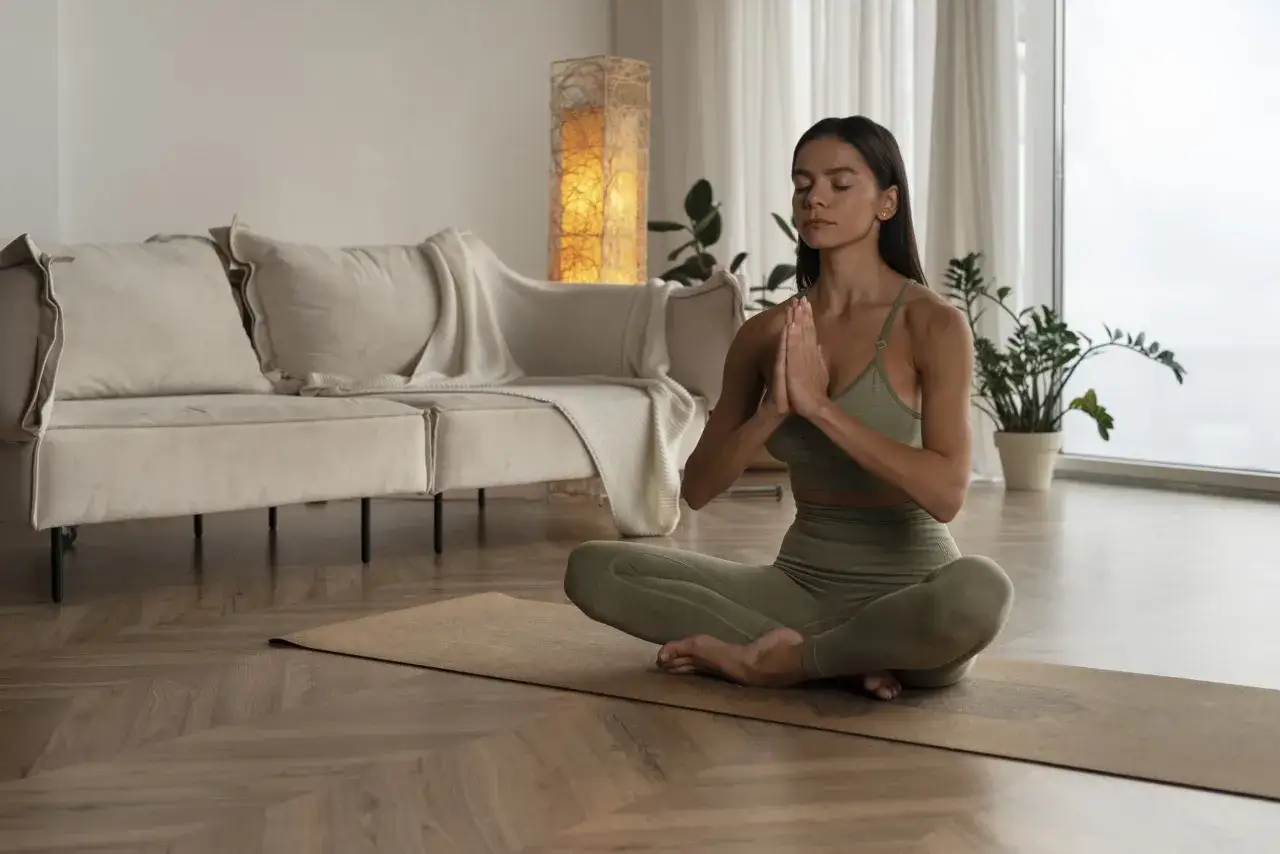 Frau meditiert auf einer Matte, um zu lernen, wie kann ich ich selbst sein. Ein gem&uuml;tliches Wohnzimmer mit Sofa und Lampe im Hintergrund.