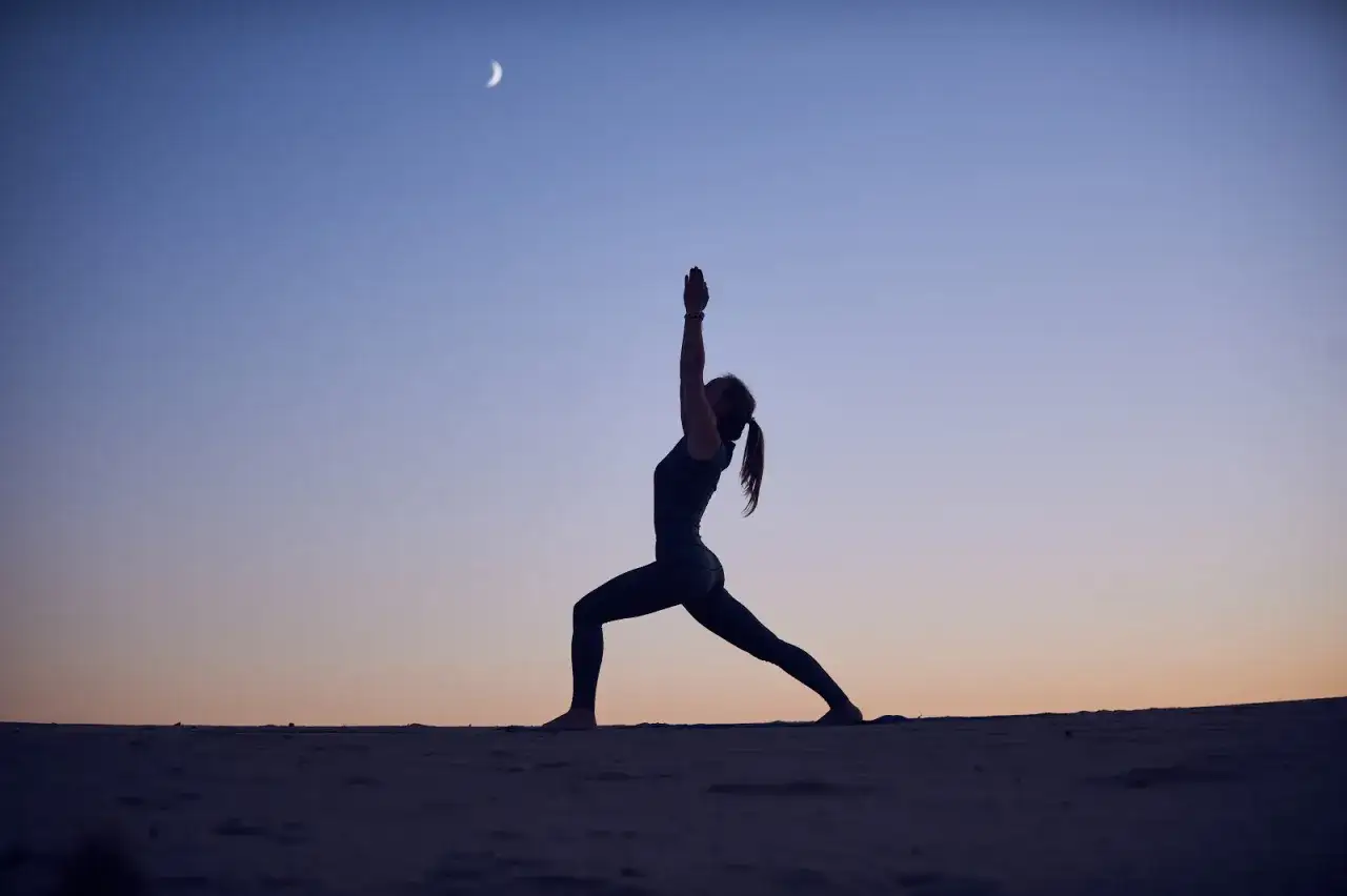 Silhouette einer Frau in Krieger-Pose bei Sonnenuntergang, der Sichelmond leuchtet am Himmel. Yoga-Mond-Moment.