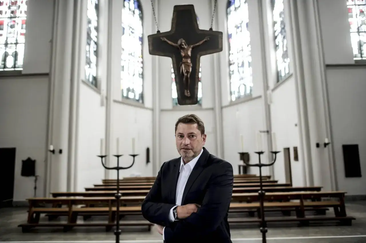 Ein Mann im Anzug steht in einer Kirche vor einem Kruzifix. Er ist ein priester im ruhestand, der &uuml;ber seine Vergangenheit nachdenkt.