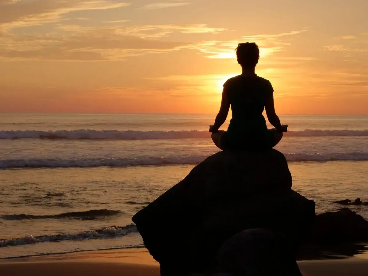 Silhouette einer Person in Lotussitz auf einem Felsen am Meer bei Sonnenuntergang. Sie meditiert und denkt: 