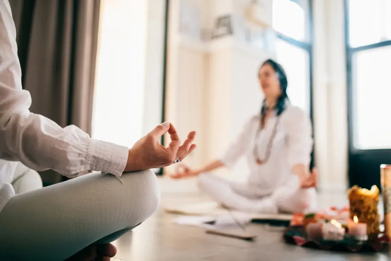 Zwei Frauen in wei&szlig;er Kleidung praktizieren aktive Meditation im Lotussitz. Kerzen und eine Ananas auf einem Tisch deuten auf eine Zeremonie hin.