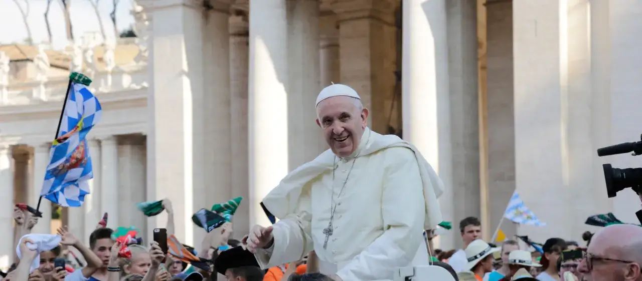 Papst Franziskus winkt einer jubelnden Menge zu. Fahnen und Handys sind erhoben, w&auml;hrend er in seinem wei&szlig;en Gewand vorbeif&auml;hrt.