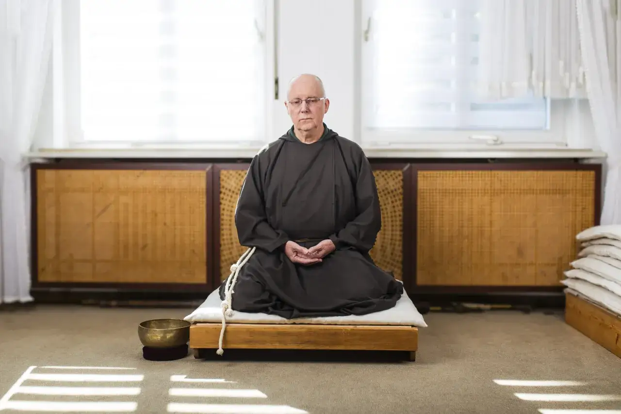Ein Mann in brauner M&ouml;nchsrobe, der an Kardinal Ratzinger erinnert, sitzt in meditativer Haltung auf einem Podest.