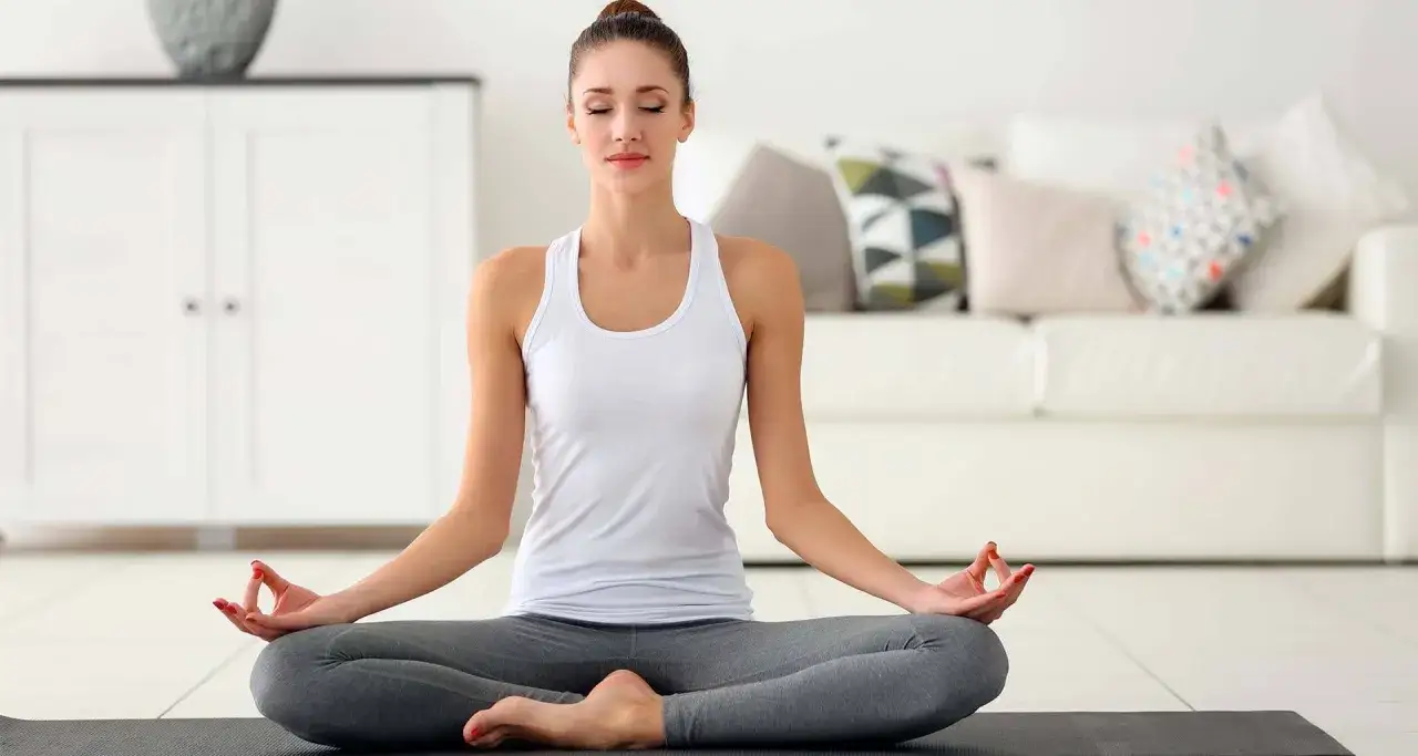 Eine junge Frau in bequemer Kleidung sitzt im Lotussitz auf einer Yogamatte. Sie schlie&szlig;t die Augen und legt ihre H&auml;nde in Mudras. Perfekt f&uuml;r yoga einsteiger.