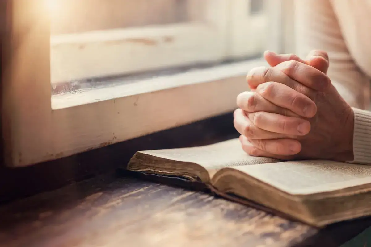 Betende H&auml;nde ruhen auf einem alten Buch am Fenster. Spirituell l&auml;sst man die Dinge fallen, wenn man sich dem Glauben zuwendet.