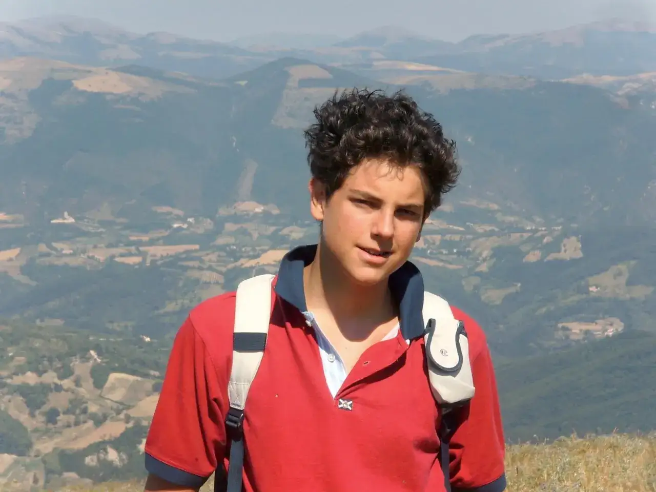 Carlo Acutis, ein junger Mann mit lockigem Haar, trägt ein rotes Poloshirt und einen Rucksack vor einer bergigen Landschaft.