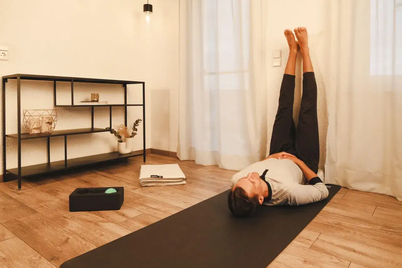 Mann in entspannter Pose mit Beinen an der Wand, Teil seiner yoga abendroutine. Eine Matte, ein Handtuch und eine Massageball-Box liegen bereit.