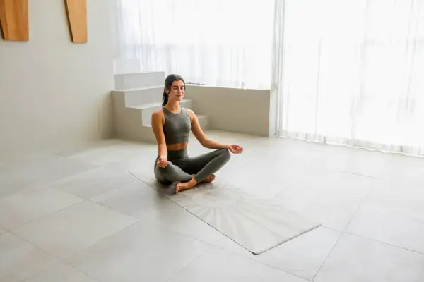 Frau in Yoga-Kleidung meditiert auf einer Matte mit einem Sonnensymbol. Der Yoga-Mantra-Text ist auf der Matte angedeutet.