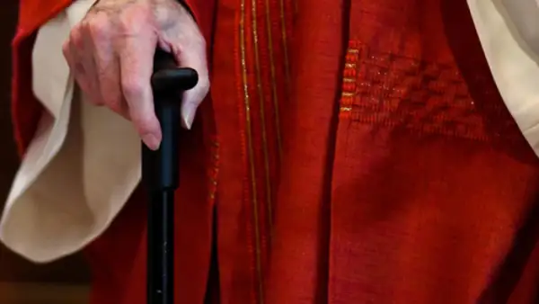 Hand eines Priesters im Ruhestand, der einen Gehstock hält. Er trägt eine rote Soutane mit goldenen Verzierungen.