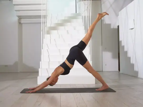 Frau in schwarzer Sportkleidung praktiziert Jivamukti Yoga in der Hundepose mit einem Bein erhoben, vor einer modernen weißen Treppe.