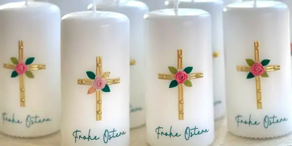 Mehrere weiße Osterkerzen mit goldenen Kreuzen, rosa Rosen und grünen Blättern. Die Osterkerze Bedeutung wird durch diese Symbole unterstrichen. "Frohe Ostern" steht geschrieben.
