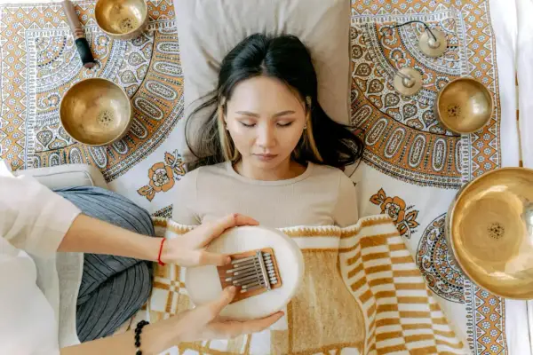 Eine Frau liegt entspannt, während eine andere Person ihr eine Kalimba über dem Bauch hält. Klangschalen und ein Stößel sind um sie herum arrangiert, was auf eine Klang Meditation hindeutet.