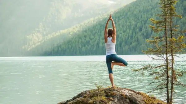 Frau in Baum-Pose am Seeufer, umgeben von Bergen. Ein friedlicher Moment des Yoga.