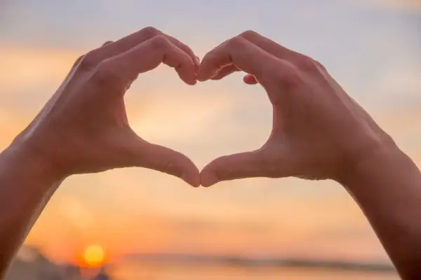 Hände formen ein Herz vor einem Sonnenuntergang. Eine spirituelle Person drückt Liebe und Verbundenheit aus.
