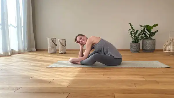 Frau in grauer Kleidung praktiziert Yoga Yin auf einer Matte. Sie lächelt entspannt und stützt ihren Kopf auf der Hand.
