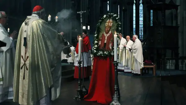 Ein Kardinal leitet eine gesungene katholische Messe. Ein Bildnis des Papstes, umrahmt von Blumen, steht im Mittelpunkt.