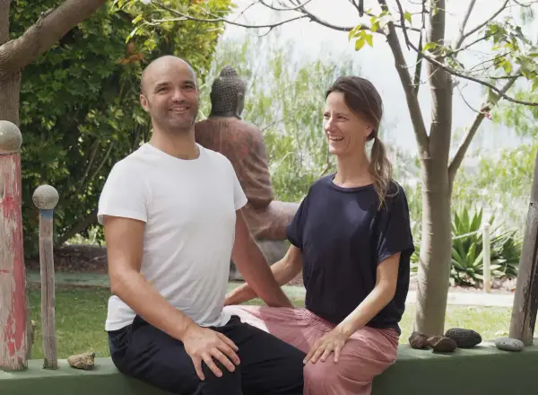 Ein Mann und eine Frau lächeln in einer grünen Umgebung. Sie sind die Gastgeber eines Ayurveda-Podcasts, der sich mit Wohlbefinden und Balance beschäftigt.