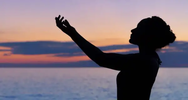Silhouette einer Frau mit erhobenen Armen vor einem farbenfrohen Himmel und Meer. Sie scheint einen **chant deutsch** zu singen.