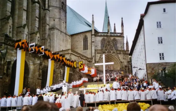 Große Menschenmenge vor einer Kathedrale, die an einem Hochamt teilnimmt. Fahnen und Girlanden schmücken die Fassade.