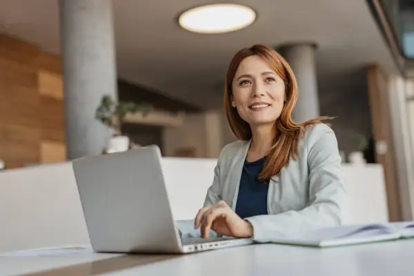 Eine Frau mit roten Haaren arbeitet an ihrem Laptop. Sie lächelt und blickt nach oben, als ob sie über ihre nächste Coaching-Sitzung nachdenkt. Ihr life coach job erfüllt sie.